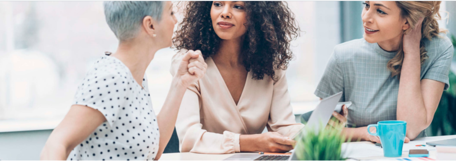 Three women in conversation during a professional meeting with one gesturing and others listening.